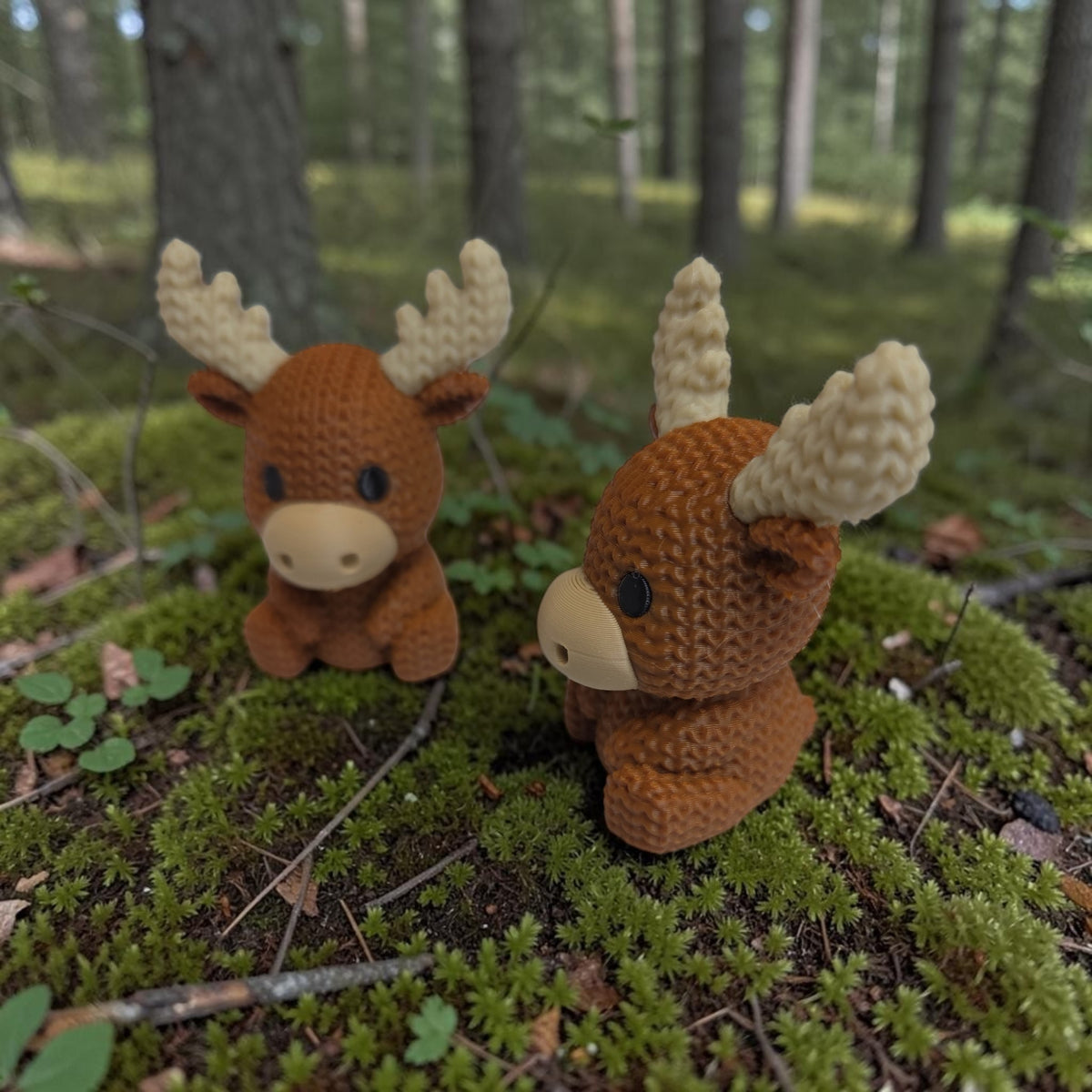 Two HPHobbies 3D printed faux knit moose figurines with brown bodies and cream antlers sitting on forest moss.